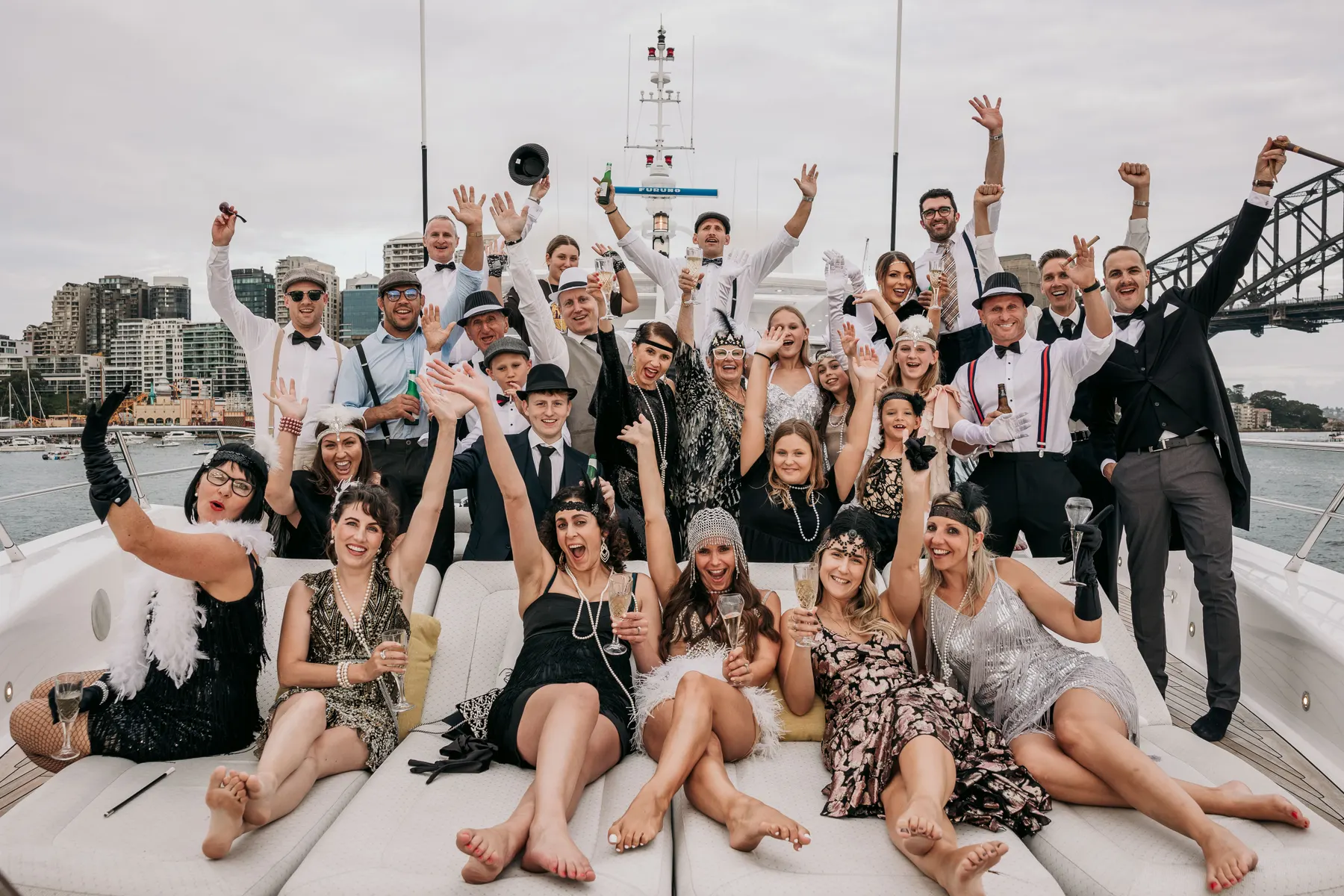 Guests in 1920s costume cheering on the foredeck of a yacht in the afternoon, the Harbour Bridge behind