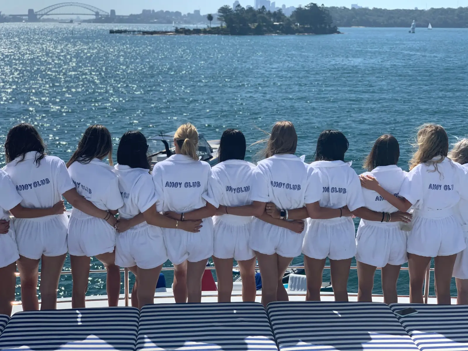 The Ahoy Club team in matching white terry jumpsuits, arm in arm on the deck of a yacht, looking across Sydney Harbour to the Harbour Bridge