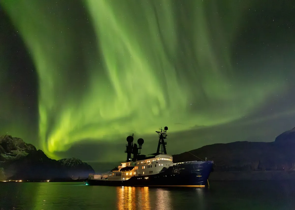 ARCTIC at anchor under the northern lights, her decks lit up and reflected in the fjord