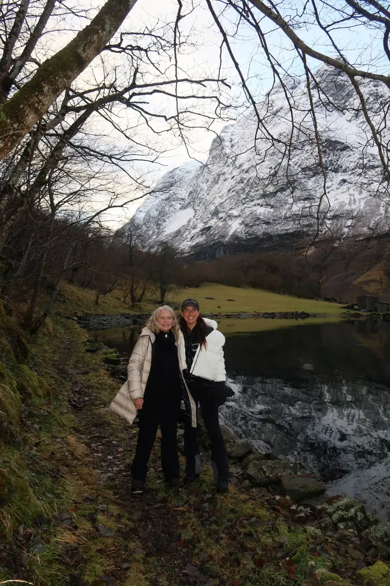 Ellie and a guest on the trail beside the fjord, a snow-covered mountain reflected in the still water